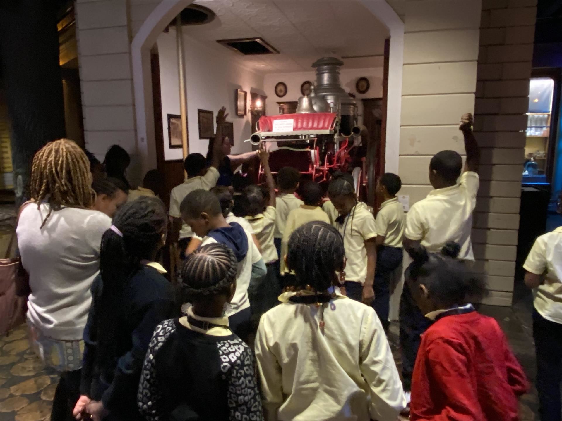 students gathered around a red antique fire truck, raising their hands to ask questions